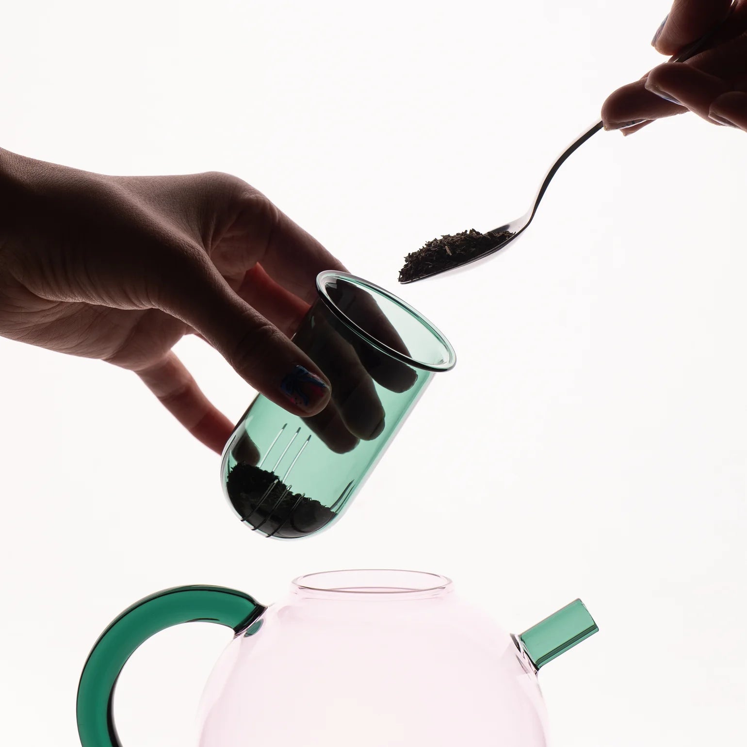 Person adding black powder from a spoon into a green glass container, with a pink glass container below.