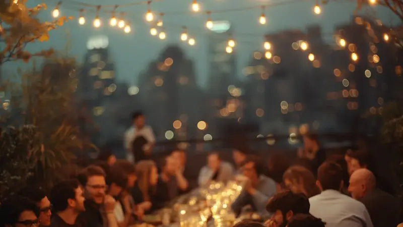People gathered around a table at night with string lights and blurred cityscape in the background